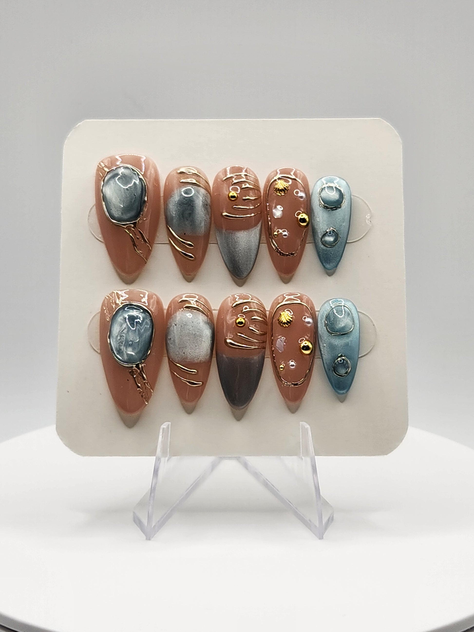 A set of 10 decorative press-on nails displayed on a white card, featuring a mix of metallic gold, dusty rose, blue, and gray designs with pearl embellishments.