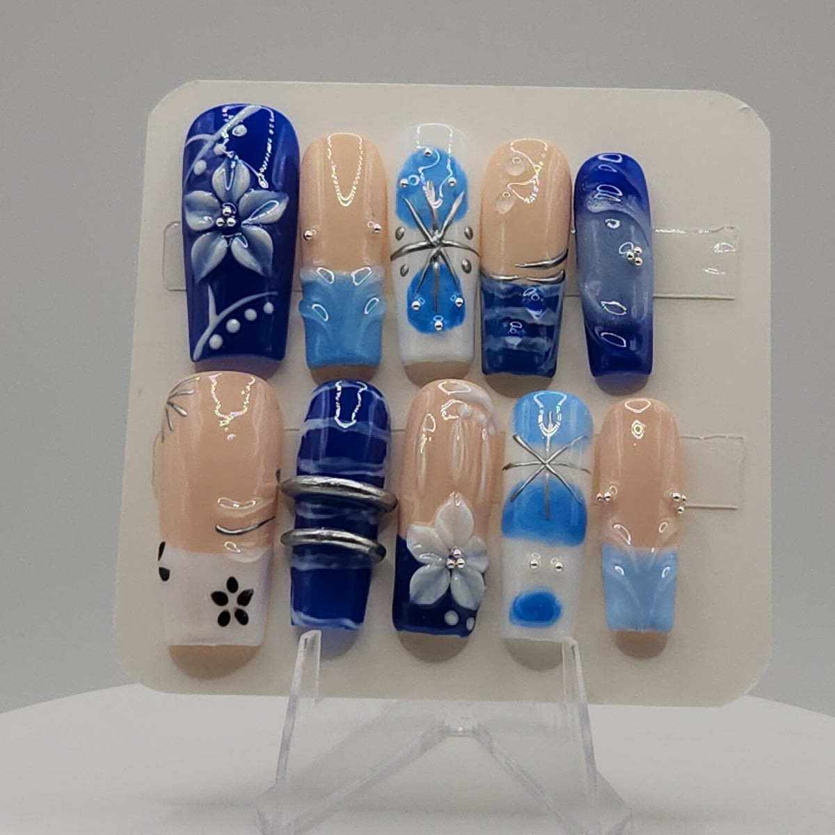 A set of 10 press-on nails displayed on a clear stand, featuring a mix of beige and blue colors with various hand-painted patterns including floral, striped, and abstract designs.