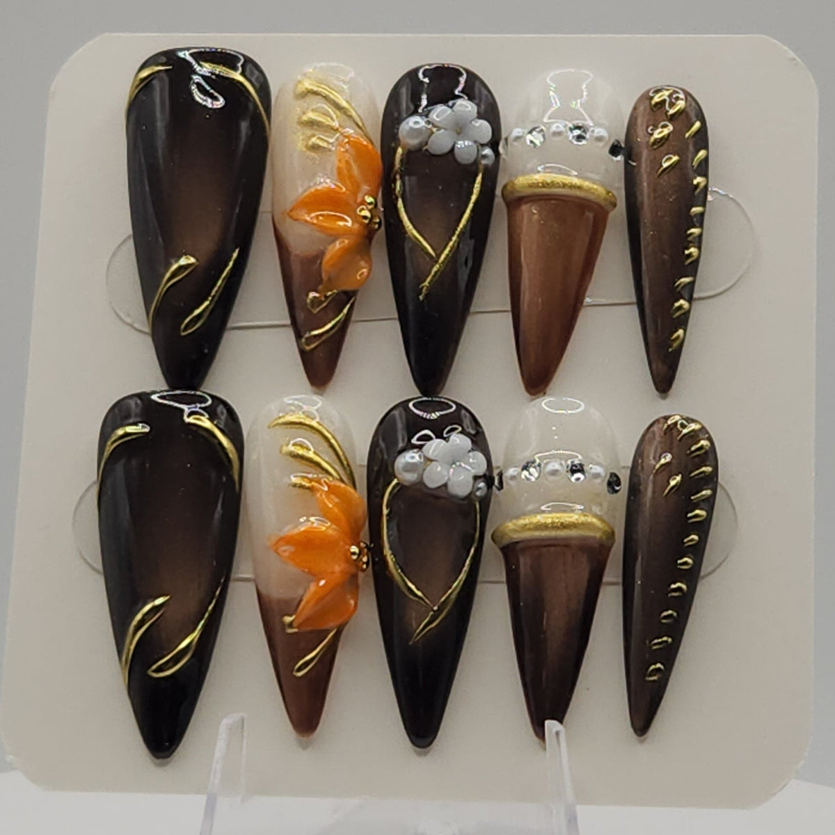 A set of 10 coffin-shaped press-on nails displayed on a white card stand. The nails feature a mix of glossy dark brown, warm brown, and black designs, decorated with gold glitter, orange floral accents, white pearl details, and small 3D elements.